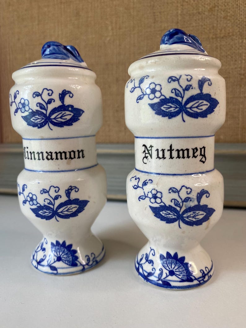 May include: Two white ceramic spice jars with blue floral designs and lids. One jar reads "Cinnamon" and the other reads "Nutmeg" in black lettering. The jars have a bulbous shape and are approximately 4 inches tall.