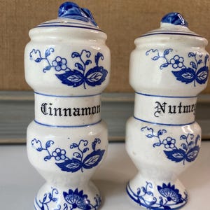 May include: Two white ceramic spice jars with blue floral designs and lids. One jar reads "Cinnamon" and the other reads "Nutmeg" in black lettering. The jars have a bulbous shape and are approximately 4 inches tall.
