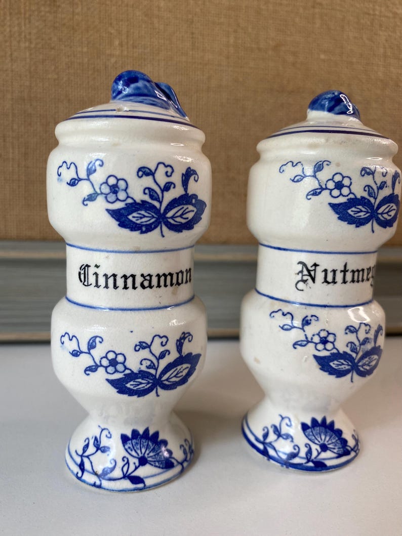 May include: Two white ceramic spice jars with blue floral designs and lids. One jar reads "Cinnamon" and the other reads "Nutmeg" in black lettering. The jars have a bulbous shape and are approximately 4 inches tall.