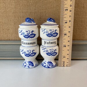 May include: Two white ceramic spice jars with blue floral designs and lids. One jar is labeled "Cinnamon" and the other "Nutmeg". Each jar is approximately 4 inches tall.