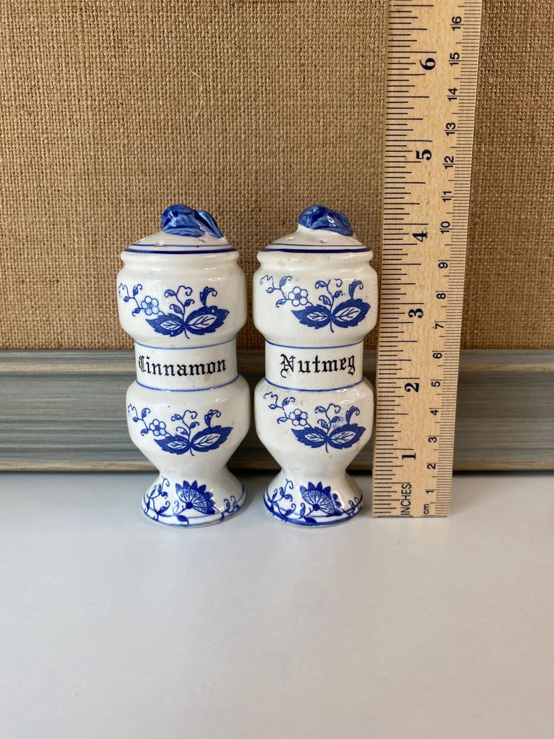May include: Two white ceramic spice jars with blue floral designs and lids. One jar is labeled "Cinnamon" and the other "Nutmeg". Each jar is approximately 4 inches tall.