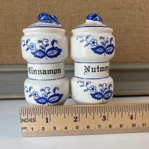 May include: Two white ceramic spice jars with blue floral designs and lids. One jar is labeled "Cinnamon" and the other "Nutmeg". A wooden ruler with inches and centimeters is in the foreground.
