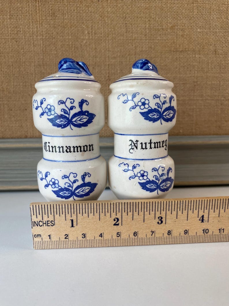 May include: Two white ceramic spice jars with blue floral designs and lids. One jar is labeled "Cinnamon" and the other "Nutmeg". A wooden ruler with inches and centimeters is in the foreground.