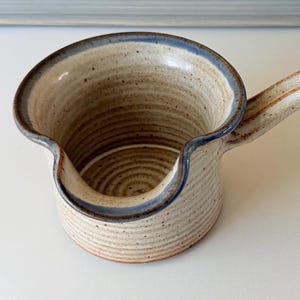 May include: A ceramic creamer with a speckled beige finish and a blue-rimmed spout. The vessel features a ribbed texture on the exterior and interior, a wide circular opening, and a single handle extending from the side, showcasing a handcrafted look.