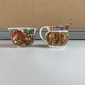 May include: A small, vintage ceramic creamer and sugar bowl set. The creamer has a handle and spout, while the sugar bowl is open. Both pieces feature a detailed landscape scene in shades of brown, green, and white, with a decorative rim.