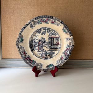 May include: A vintage decorative plate with a cream-colored background and a detailed blue and white Asian-inspired scene. The plate features a floral border with pink, blue, and green accents. It is displayed on a small red stand.