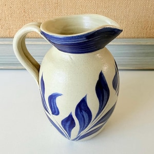 May include: A cream-colored ceramic pitcher featuring a hand-painted dark blue leaf pattern around the body and a solid dark blue rim. The pitcher has a curved handle on the left side and a wide pouring spout, showcasing a smooth, glazed finish.