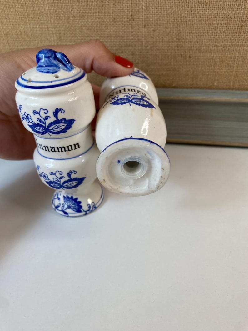 May include: White ceramic spice shakers with blue floral designs and the words "cinnamon" and "nutmeg". The shakers have a classic, vintage aesthetic, with a small handle on the lid.