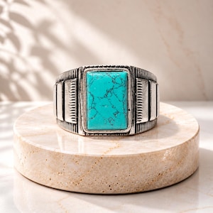 May include: A silver-toned ring featuring a rectangular, bright turquoise stone with dark veining. The band has a textured, oxidized finish with vertical grooved detailing on both sides of the central stone setting.