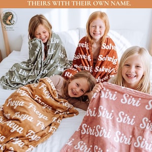 Puede incluir: Cuatro mantas suaves en verde salvia, naranja óxido, marrón dorado y rosa empolvado, cada una con un estampado de nombre blanco repetido. Las mantas cubren a cuatro niños sentados en una cama. El texto superior dice: ONE WONDERFUL GIFT IDEA FOR ALL THE GRANDKIDS. SAME COZY WARMTH, UNIQUELY THEIRS WITH THEIR OWN NAME.