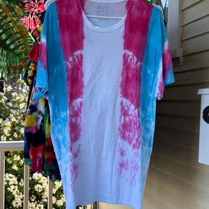 Peut inclure: Un t-shirt blanc avec un motif tie-dye. Le t-shirt présente des rayures verticales de teinture rose et bleue. D'autres t-shirts tie-dye sont visibles en arrière-plan, suspendus à un portant. Le t-shirt est suspendu à un cintre blanc.
