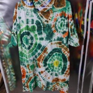 May include: A tie-dye t-shirt in shades of green, brown, orange, and yellow. The shirt has a spiral pattern and is labeled "XLT". The shirt is hanging on a white rack.
