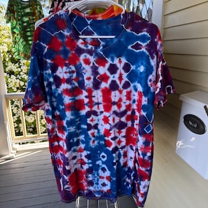 Peut inclure: Un t-shirt tie-dye à col en V aux couleurs rouge, blanc et bleu, suspendu à un cintre blanc. Le t-shirt présente un motif en losange et est exposé à l'extérieur. D'autres t-shirts tie-dye sont visibles en arrière-plan.