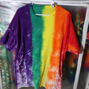 Peut inclure: T-shirt tie-dye XL avec un motif arc-en-ciel. Le t-shirt présente des rayures verticales violettes, vertes, jaunes, oranges et rouges. Le vêtement est suspendu à un cintre blanc.