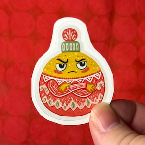 May include: A sticker featuring a cartoon Christmas ornament with an angry face. The ornament is yellow with a red and white patterned design. The sticker is held against a red background.