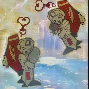 May include: Two cartoon character keyrings. The characters are white with red and black accents, and have red wings. The keyrings are on a holographic background. One keyring is holding a grey object. The keyrings are attached to heart-shaped clasps.