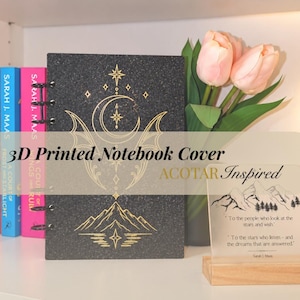 Pode incluir: Uma capa de caderno impressa em 3D, preta e brilhante, com desenhos de linhas douradas de uma lua crescente, estrelas e picos de montanhas. A capa inclui uma encadernação de argolas e está ao lado de uma pequena placa decorativa.