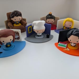 Puede incluir: Colección de seis figuras estilizadas, cada una representando un personaje de la serie de televisión "Friends". Las figuras están sobre bases coloridas, con accesorios en miniatura como una mesa de café, una caja de pizza y un sillón. Las figuras son de estilo caricatura.