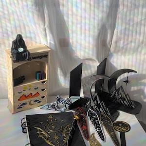 Pode incluir: Um conjunto de artigos de papelaria incluindo um caderno de espiral preto com gráficos celestiais dourados, uma caneta a condizer, vários marcadores decorativos, uma pequena prateleira de madeira com gavetas pintadas e suportes para livros em metal preto em forma de lua.