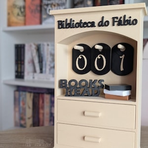 Può includere: Una libreria in legno beige con la scritta "Biblioteca do Fábio" e "BOOKS READ". Ha tre etichette nere con i numeri "001". La libreria ha due cassetti e una piccola mensola con tre piccoli blocchi decorativi.
