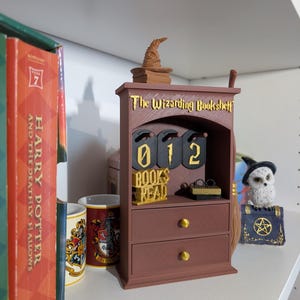 Pode incluir: Um expositor castanho "The Wizarding Bookshelf" com um chapéu seletor e uma vassoura. O expositor tem uma gaveta e um contador mostrando "012 BOOKS READ". Um livro de Harry Potter e uma caneca da Grifinória estão na prateleira.