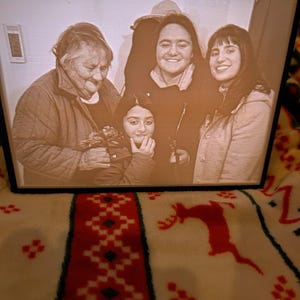 Pode incluir: Fotografia em tons sépia emoldurada com quatro pessoas sorrindo. Os sujeitos incluem uma mulher mais velha, uma menina e duas outras mulheres. A moldura é preta e a fotografia está sobre um cobertor branco com um padrão vermelho.