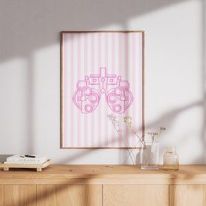 Könnte beinhalten: Ein gerahmter Druck mit einer rosa Umrisszeichnung eines Phoropters auf einem rosa-weiß gestreiften Hintergrund. Der Druck wird auf einem Holzschrank mit einer Vase und anderen dekorativen Gegenständen ausgestellt.