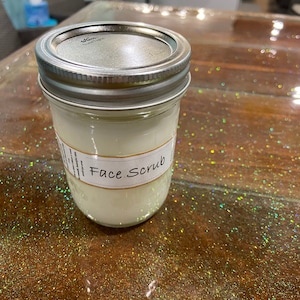 May include: A clear glass jar with a silver lid filled with a white face scrub. The jar has a white label that says "Face Scrub".