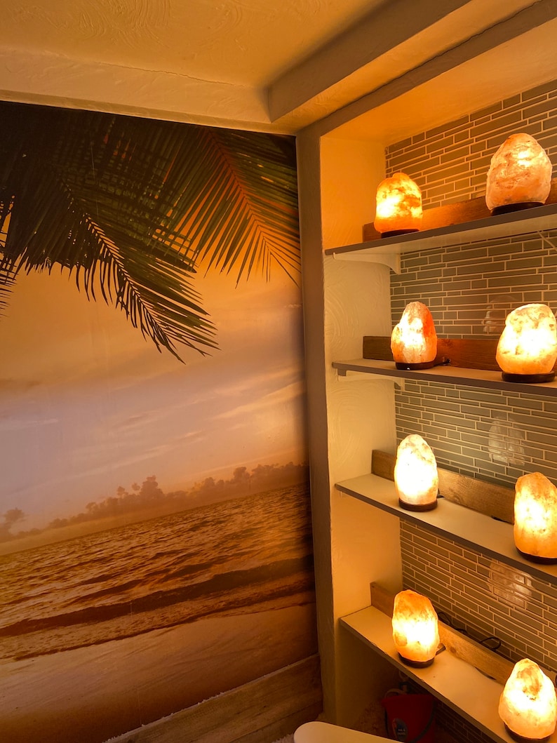 May include: A room with a tropical beach mural and several illuminated Himalayan salt lamps on shelves. The lamps emit a warm, orange glow, creating a relaxing ambiance. The wall behind the shelves has a gray tile design.
