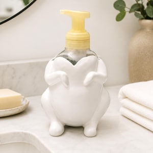 May include: A white ceramic soap dispenser shaped like a whole roasted chicken with a yellow plastic pump mechanism. The dispenser features a rounded body with sculpted legs and wings, holding a clear plastic bottle for liquid soap inside.