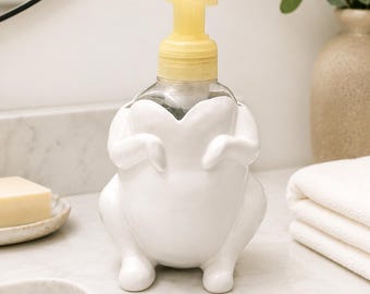 Peut inclure: Un distributeur de savon en céramique blanche en forme de poulet rôti entier avec un mécanisme de pompe en plastique jaune. Le distributeur présente un corps arrondi avec des pattes et des ailes sculptées, contenant une bouteille en plastique.