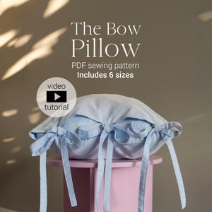 Könnte beinhalten: Ein hellblaues Kissen mit Schleifen und langen Bändern, das auf einem rosa Hocker liegt. Das Kissen hat ein Schleifendesign an den Seiten. Der Text auf dem Bild lautet "The Bow Pillow" und "PDF sewing pattern Includes 6 sizes". Ein Video-Tutorial-Symbol ist ebenfalls vorhanden.