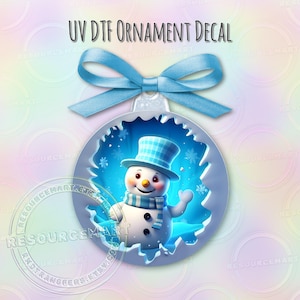 May include: A UV DTF ornament decal featuring a smiling snowman wearing a blue plaid top hat and scarf. The snowman is inside a blue, circular frame with snowflakes. A light blue ribbon is attached to the top of the ornament.