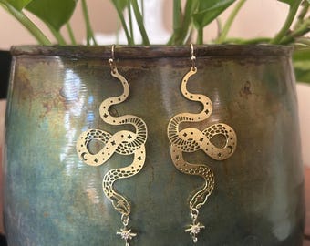 May include: A pair of gold-colored snake earrings. Each earring features a stylized snake design with etched details, including stars and crescent moons. A small star-shaped charm with a crystal hangs from the bottom of each earring. The earrings are displayed against a green and brown background.