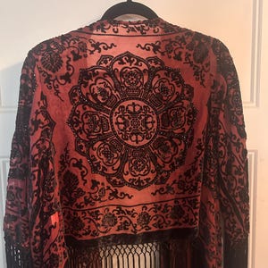 May include: A red and black kimono-style jacket featuring a prominent black velvet floral and mandala-inspired pattern across the back. The garment is accented with intricate black beadwork and finished with long black fringe along the bottom hem.