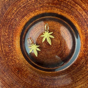 Puede incluir: Un par de pendientes dorados, cada uno con un diseño detallado de hoja de cannabis. Los pendientes se muestran sobre una superficie marrón y texturizada, creando una estética cálida y terrosa. Los pendientes son pequeños, con cierre de aro.