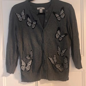 May include: A charcoal grey button-down cardigan featuring long sleeves and a crew neckline. The garment is decorated with several embroidered butterfly motifs in black and white thread. A Bass brand label is visible at the inner collar.