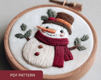 May include: Embroidered snowman in a wooden hoop. The snowman has a carrot nose, a claret scarf, and a brown hat with an orange band. Green leaves and the text "PDF PATTERN" are also visible.