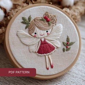 May include: Hand-stitched fairy embroidery in a wooden hoop. The fairy is depicted with a red dress, white wings, and brown hair adorned with red berries. The design includes green leaves and the text "PDF PATTERN" at the bottom.