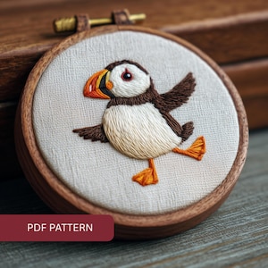 May include: Embroidered puffin bird in a wooden hoop. The bird has a white chest, brown wings, and orange feet and beak. The background is a light beige fabric. The text "PDF PATTERN" is in a red box at the bottom.