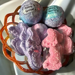 Może przedstawiać: Ozdobny pomarańczowo-brązowy kosz wypełniony produktami do kąpieli. Dwie kuliste kule do kąpieli, z napisem "Blueberry Peppermint", znajdują się nad kilkoma fioletowymi i różowymi musującymi płatkami do kąpieli w kształcie stóp, wszystkie zapakowane w przezroczysty plastik.