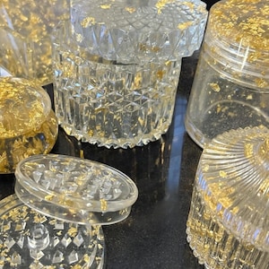 Könnte beinhalten: Klare Glasbehälter und -gläser mit Goldblatt-Akzenten. Die Gläser sind in verschiedenen Größen und Formen erhältlich, einige mit Deckeln. Das Goldblatt ist in das Glas eingebettet und erzeugt einen luxuriösen und dekorativen Effekt.