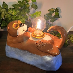 May include: A whimsical otter-shaped lamp with a glowing lightbulb. The ceramic lamp features brown and tan fur, a blue base, and a gold-colored light fixture. The otter is lying on its back, holding the lightbulb.