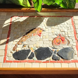 Puede incluir: Bandeja de madera para servir con diseño de mosaico. El mosaico presenta dos pájaros, uno posado y otro redondo, con colores rojo, naranja y marrón. La bandeja tiene un fondo de baldosas de color claro y un borde rojo.