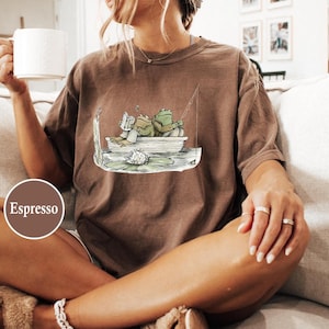 May include: Espresso-colored t-shirt featuring a whimsical illustration of two frogs in a boat. One frog reads a book, while the other fishes. The design includes a lily pad and fishing rod. The shirt is a relaxed fit.