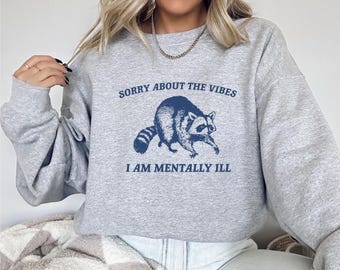 May include: A light gray sweatshirt with the text "SORRY ABOUT THE VIBES I AM MENTALLY ILL" in navy blue. A navy blue raccoon graphic is centered below the text. The sweatshirt is a casual style.