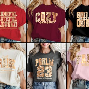 May include: Collection of six t-shirts in various colors, each featuring a different inspirational message in a decorative font. The shirts include phrases like "Thankful, Grateful, Blessed" and "God is Good."