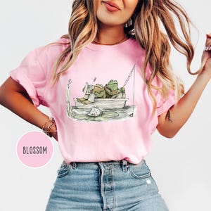 Op de afbeelding: Roze T-shirt met een grillige illustratie van twee kikkers in een boot. E&eacute;n kikker leest een boek, de ander vist. Het shirt heeft een ronde hals en korte mouwen. Het woord "BLOSSOM" staat in een roze cirkel.