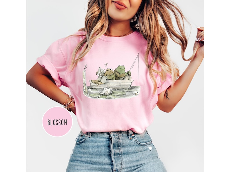 Op de afbeelding: Roze T-shirt met een grillige illustratie van twee kikkers in een boot. E&eacute;n kikker leest een boek, de ander vist. Het shirt heeft een ronde hals en korte mouwen. Het woord "BLOSSOM" staat in een roze cirkel.