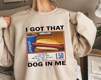 May include: Beige sweatshirt with the text "I GOT THAT DOG IN ME" and a graphic of a hot dog, soda, and price tag. The graphic includes the text "Kirkland Signature" and "1/4 POUND PLUS ALL BEEF HOT DOG AND 20 oz. SODA (With Refill)".
